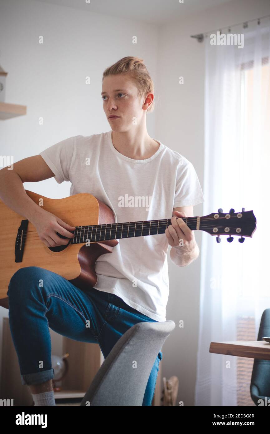 Ritratto candida di un giovane artista nel soggiorno. Il cantante tiene la sua chitarra e strums una canzone. Quarantena domestica. Realizzare il vostro sogno. In un whit Foto Stock
