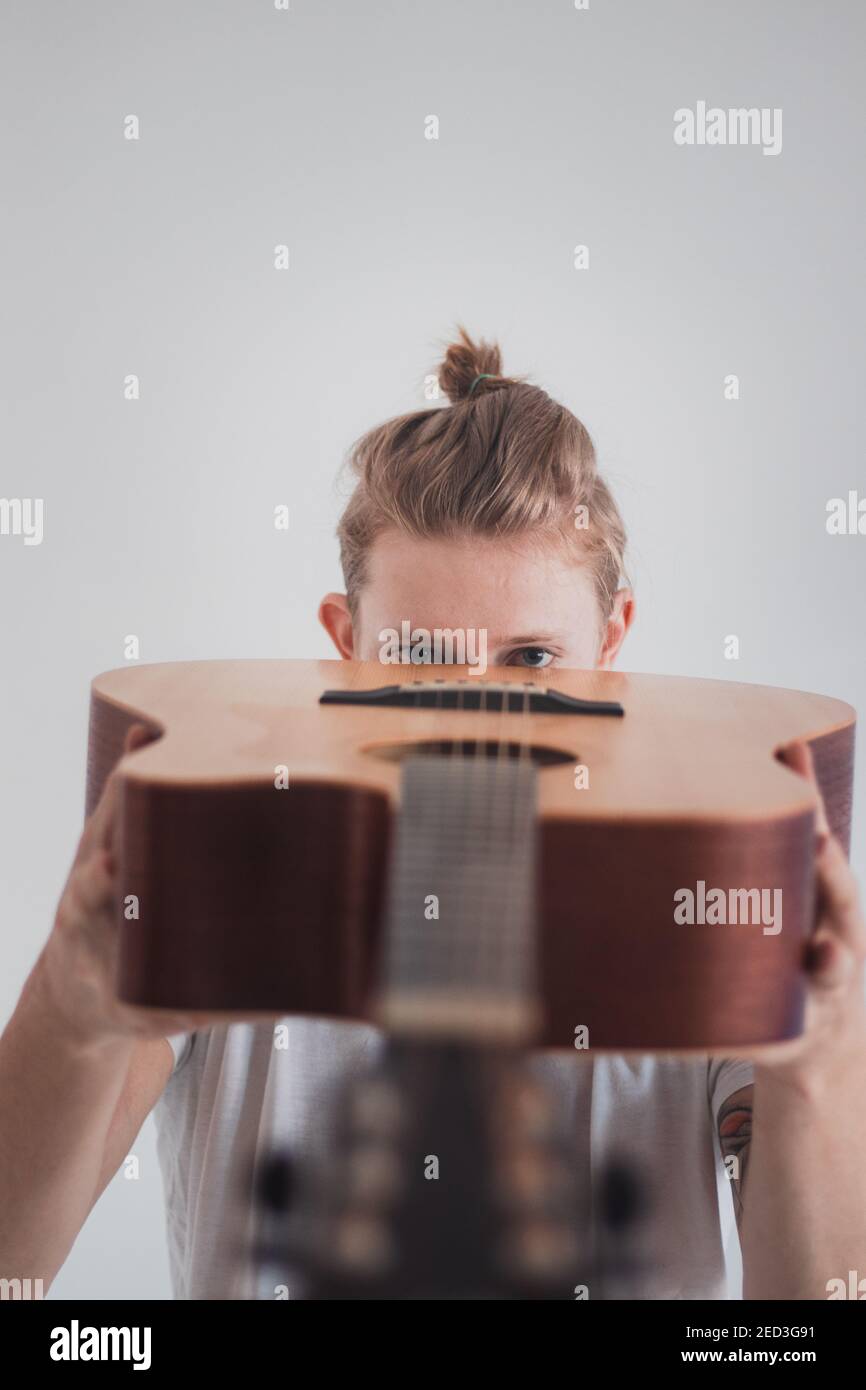 il giovane biondo guarda attraverso il suo strumento artistico - una chitarra di legno. Sfondo bianco. Ritratto binario di un uomo che canta per gioia. Quarantena domestica. Foto Stock