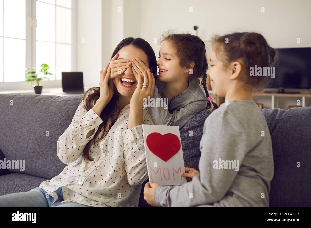 Due sorelle gemelle carine chiudono gli occhi della loro madre quando le danno una scheda fatta a mano. Foto Stock