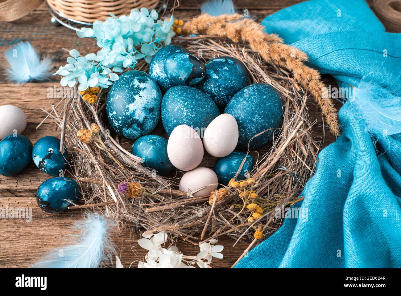 Uova di Pasqua blu-marmo in un nido di vimini con piume e fiori accanto a un tovagliolo blu e un cestino di uova pastello. Foto Stock