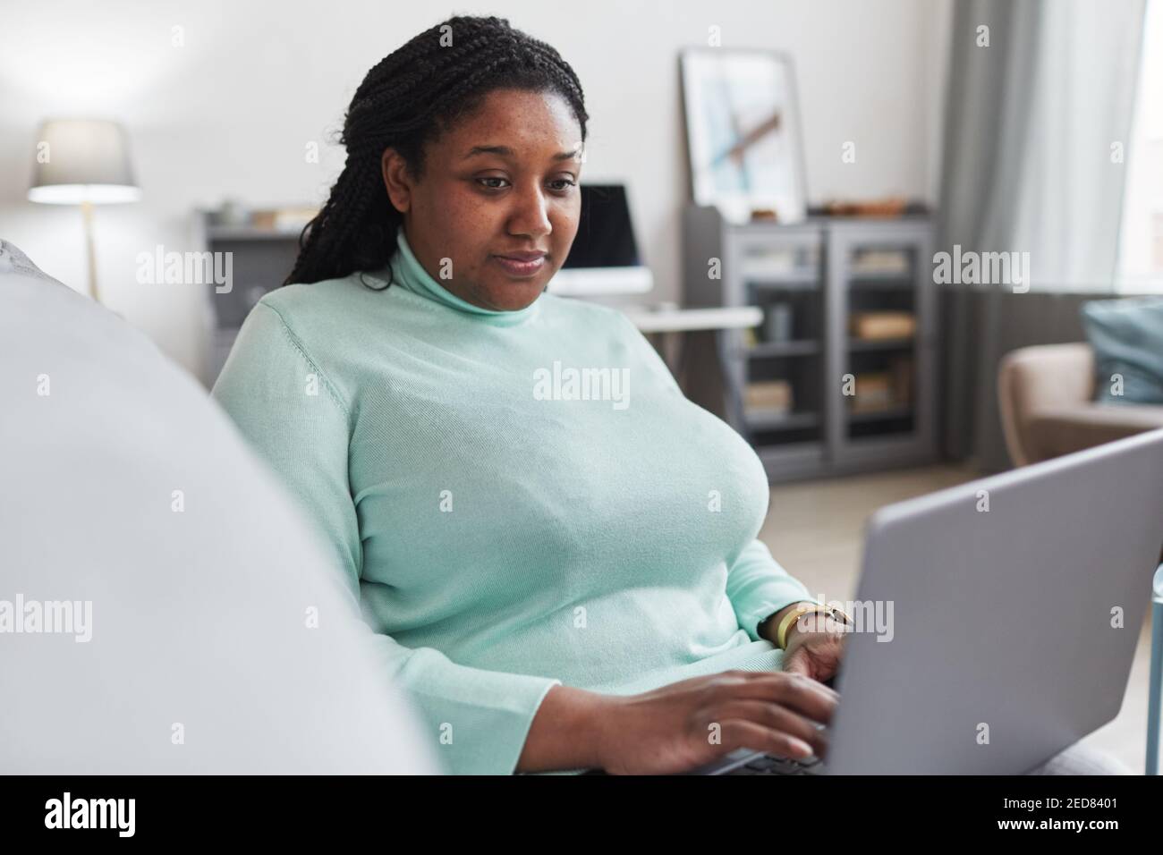 Ritratto di donna afro-americana curvy utilizzando il computer portatile mentre ti godi lavorate da casa rilassandovi sul divano in interni moderni e minimalisti Foto Stock