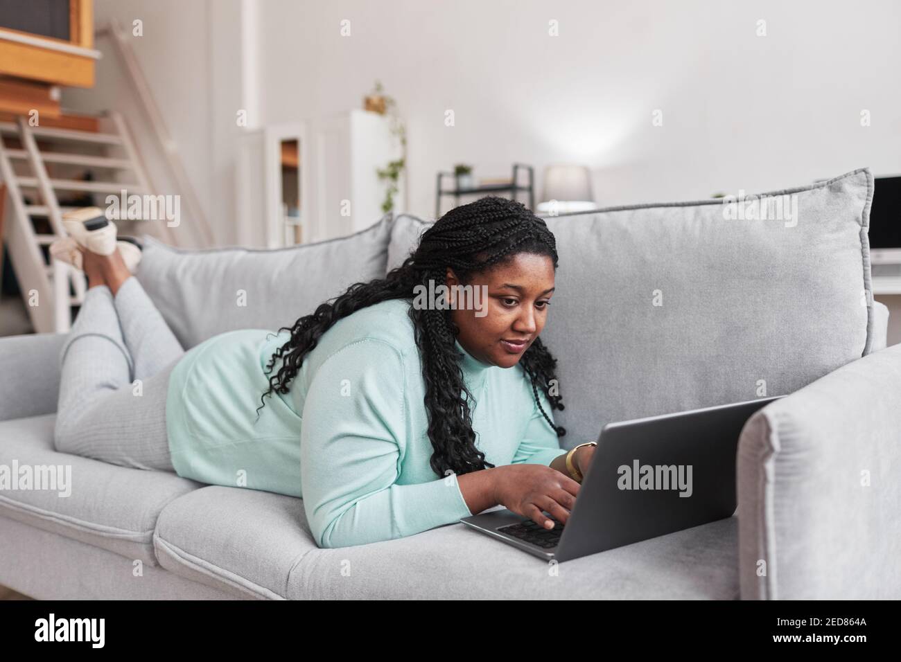 Ritratto a lunghezza intera di donna afro-americana curvy usando il laptop mentre si sdraiano sul divano e si rilassano a casa in minimal interno Foto Stock