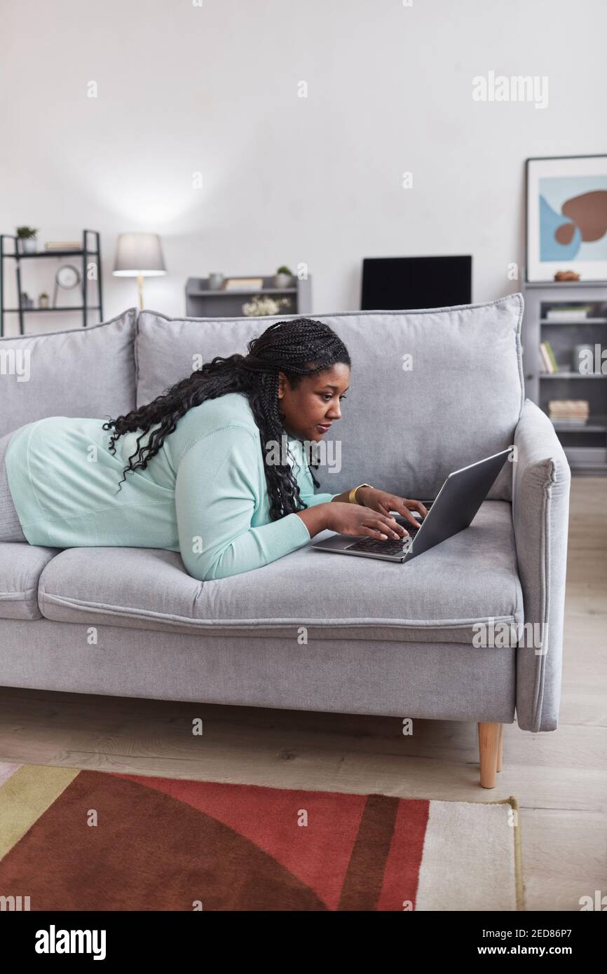 Ritratto verticale di una donna afro-americana curvilinea che usa il computer portatile mentre si sdraia sul divano e si rilassa a casa in uno spazio interno minimo, copia Foto Stock