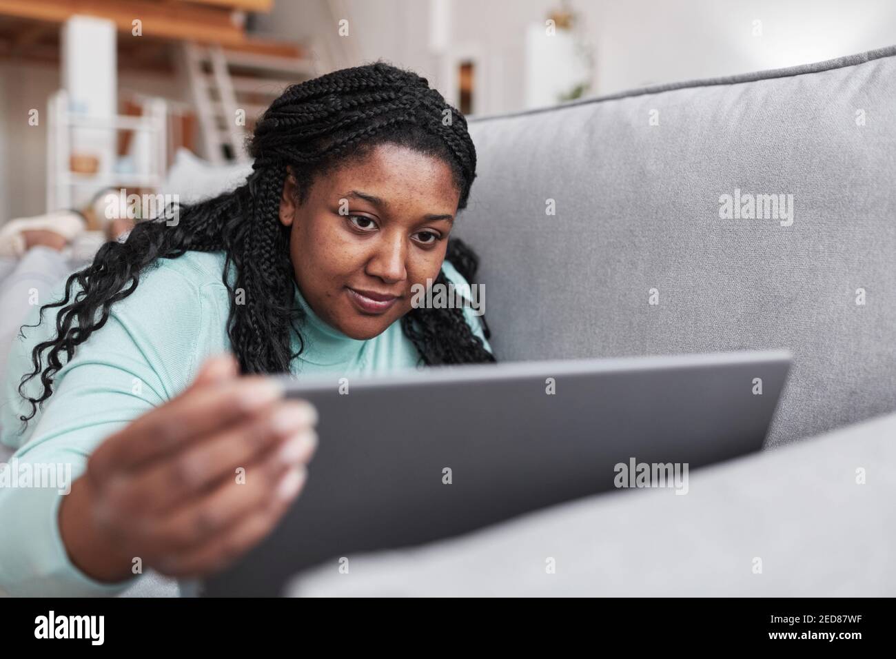 Ritratto di donna afro-americana curvy utilizzando il computer portatile mentre si sdraia sul divano e rilassarsi a casa in interni minimal, spazio di copia Foto Stock
