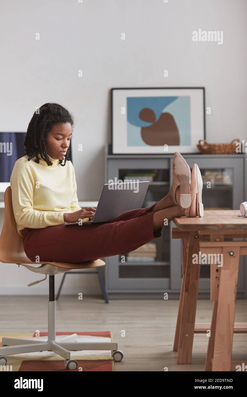 Verticale ritratto a tutta lunghezza di giovane donna afroamericana usando il laptop a casa con i piedi sul tavolo, spazio di copia Foto Stock
