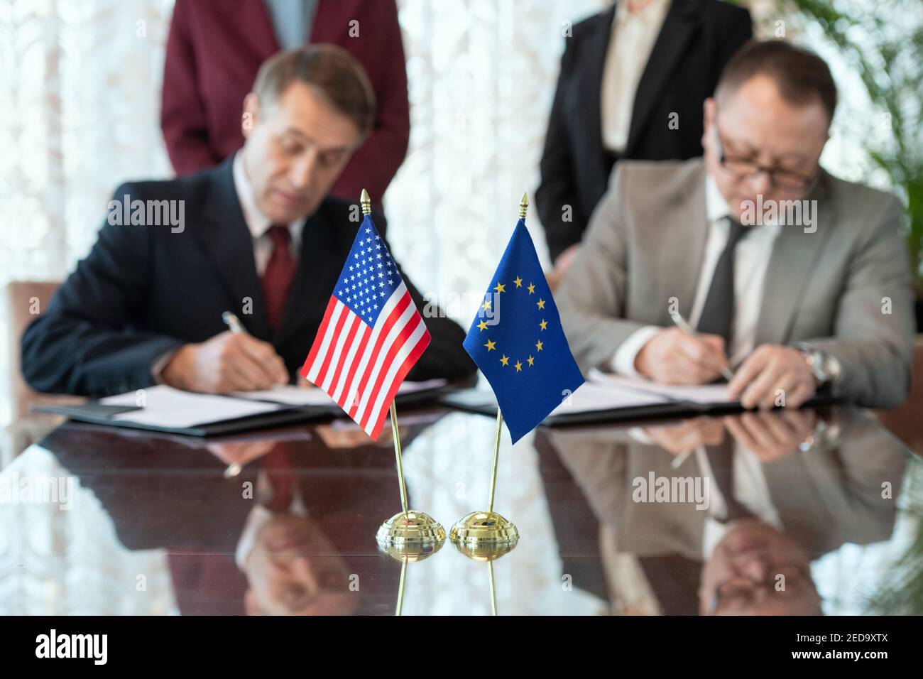Bandiere degli Stati Uniti e dell'Unione europea sul tavolo contro due partecipanti firmano il contratto dopo la negoziazione e la conclusione dell'accordo Foto Stock