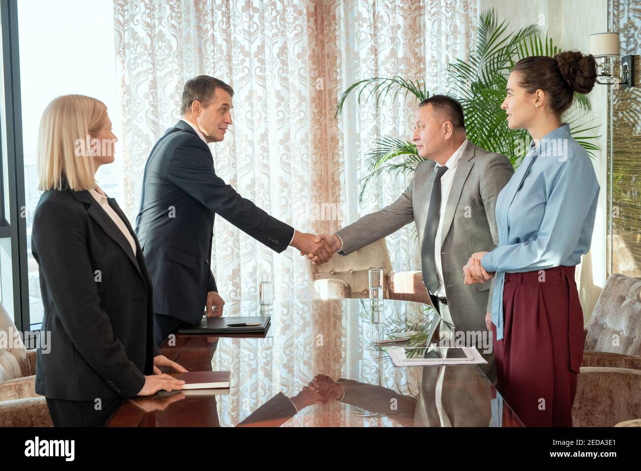 Uomini d'affari maturi di successo o delegati in formalwear salutarsi l'un l'altro con handshake sopra il tavolo dopo la negoziazione e la conclusione di un accordo Foto Stock