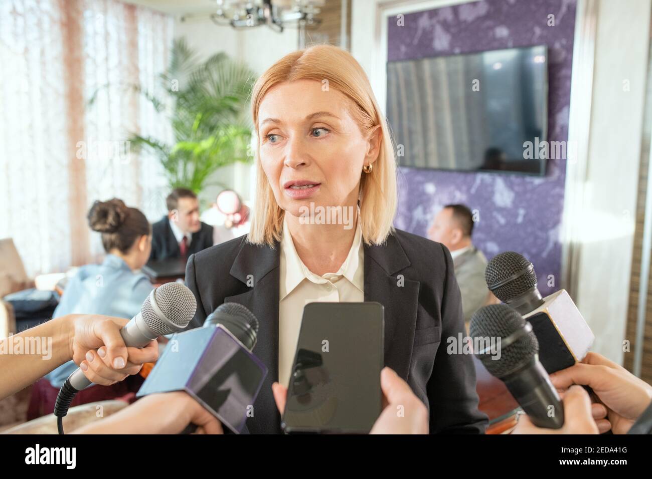 Delegato femminile biondo maturo in formalwear in piedi di fronte giornalisti con microfoni e rispondendo alle loro domande durante l'intervista Foto Stock