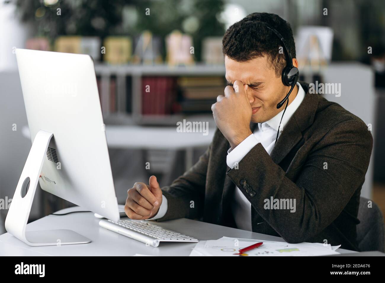 Il manager ispanico maschile o l'operatore del call center si siede sul posto di lavoro con le cuffie, utilizzando il computer. Un dipendente stanco o un tutor lavora senza pause, ha mal di testa, ha bisogno di riposo o pausa Foto Stock