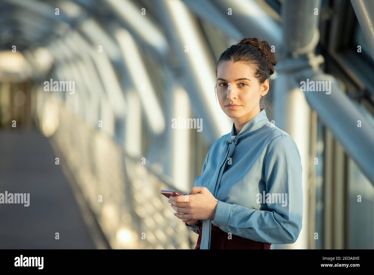 Giovane donna di brunette delegato che ti guarda mentre si scorre dentro telefono cellulare di fronte alla fotocamera all'interno di un moderno business center Foto Stock