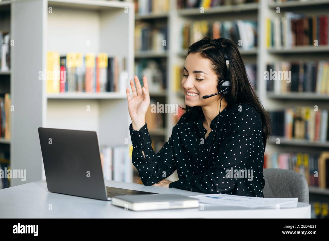 Un'attraente dipendente femminile si siede alla scrivania, facendo una videoconferenza con colleghi o clienti. Giovane donna caucasica che indossa cuffie, funziona come operatore di call center, concetto di comunicazione Foto Stock