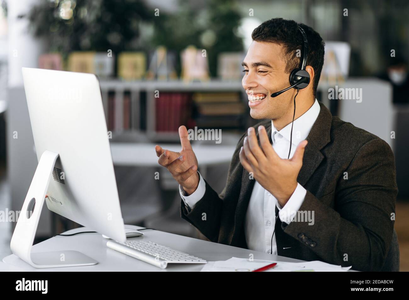 Leader, manager o tutor ispanici di successo, seduti alla scrivania indossando le cuffie, comunicano e sorridono durante una videoconferenza con colleghi o studenti Foto Stock