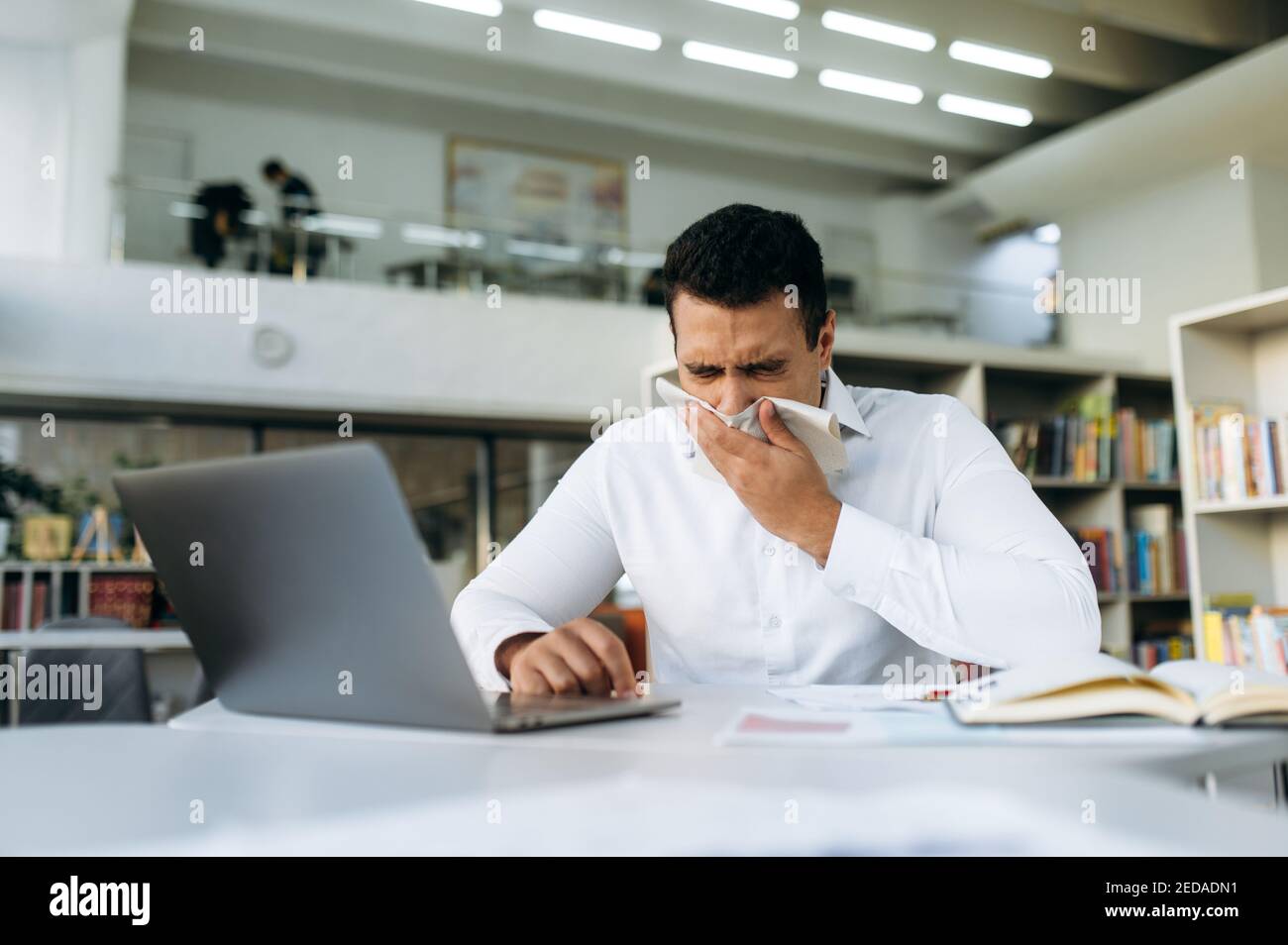 L'uomo d'affari ispanico starnutisce, usando un tissue di carta, ritenendosi malato. Manager malsano che lotta con il naso che corre, prendere un'influenza, lavorare a distanza o studiare, mentre la malattia Foto Stock