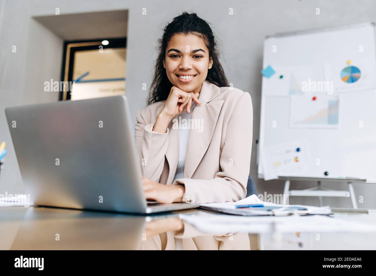 Un'elegante donna d'affari si siede alla scrivania, sorride e guarda la macchina fotografica. Una donna afroamericana che usa il laptop, sviluppa un nuovo progetto. Bella giovane donna adulta che studia o lavora a distanza Foto Stock