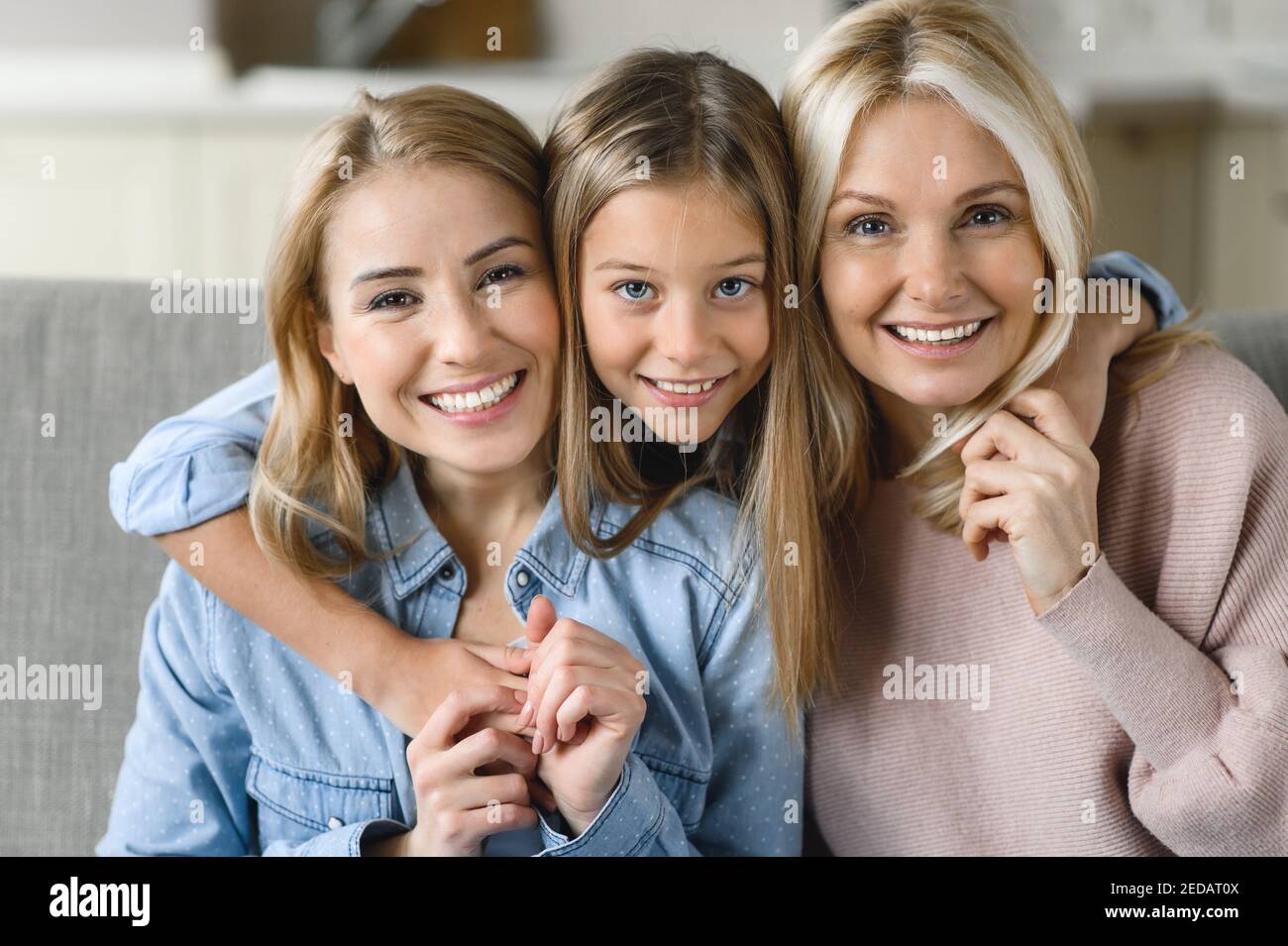 Tre generazioni femminili. Felice nonna caucasica, figlia e nipote sono seduti a casa sul divano, abbracciando, guardando la macchina fotografica e sorridendo. Rapporti e valori familiari Foto Stock