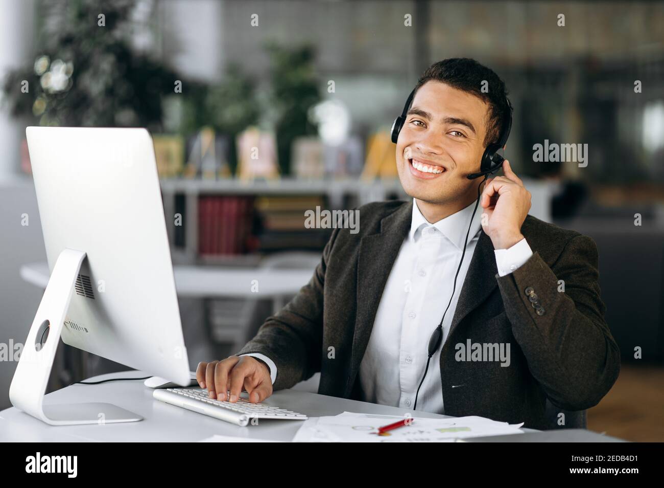 Giovane adulto maschio sta consultando i clienti, lavoro nel servizio di supporto. Felice uomo ispanico seduto alla scrivania, indossando le cuffie, funziona come operatore di call center, comunicando con i clienti Foto Stock