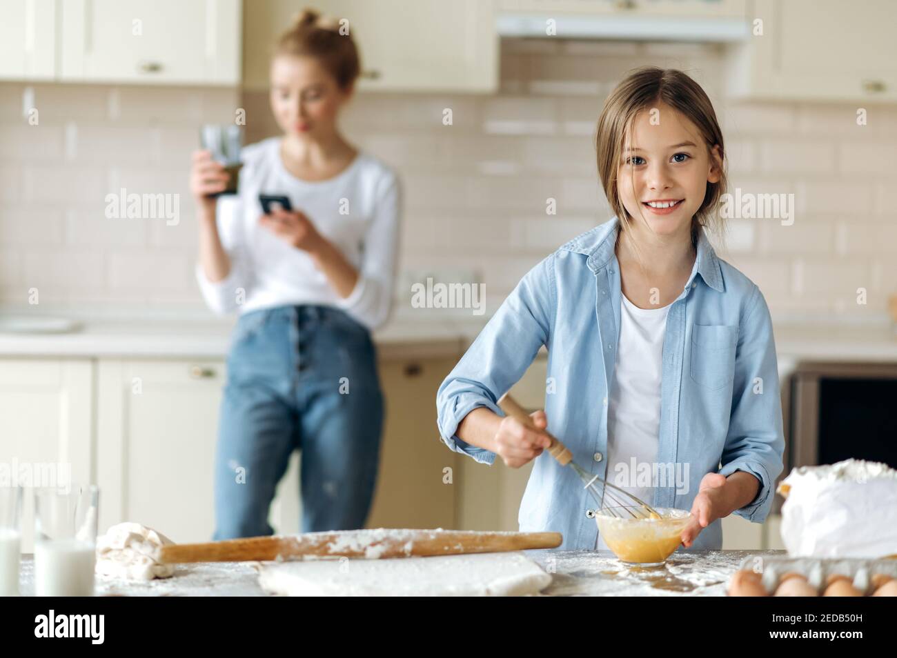 Il piccolo aiutante della mamma. Una bambina caucasica carina impara a cucinare una torta, batte le uova, guarda la macchina fotografica e sorride, mamma si alza sullo sfondo utilizzando il telefono Foto Stock