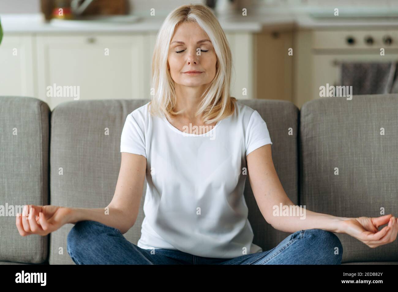 Calma attraente donna di mezza età si siede sul divano a casa e freddo. La donna anziana è pratica yoga e meditazione in posizione di loto con gli occhi chiusi, alleviare lo stress, concetto di calma Foto Stock