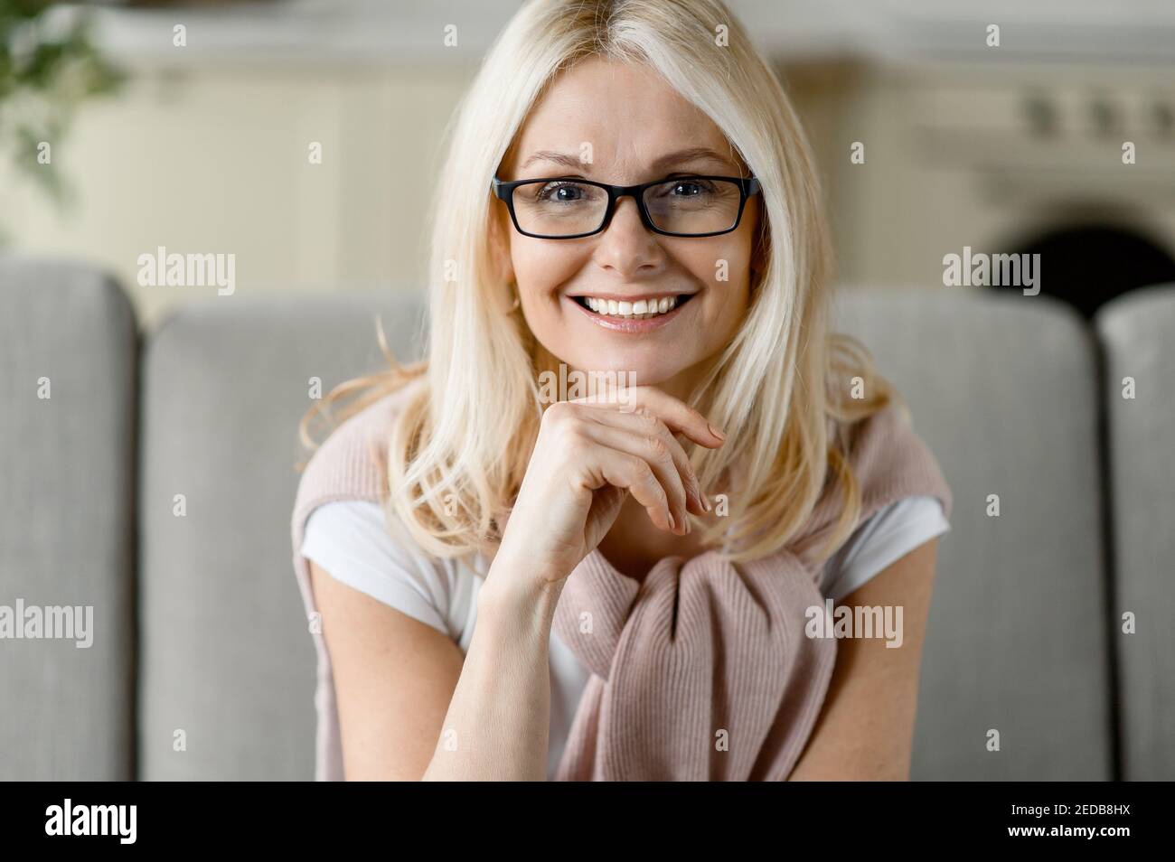 Primo piano ritratto di una donna bionda caucasica di mezza età che indossa occhiali, seduto sul divano, guardando direttamente la fotocamera con un sorriso amichevole Foto Stock