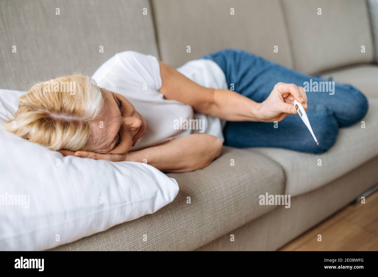 Donna malsana matura sdraiata su divano con mal di testa guarda il termometro, ha una febbre alta, virus, freddo, malattia Foto Stock