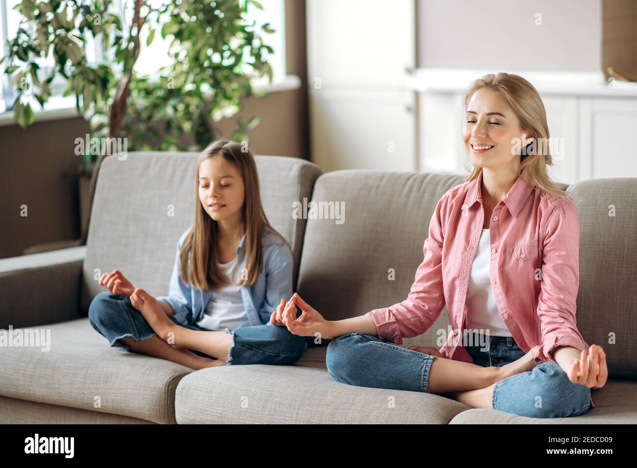 Relazione tra mamma e figlia. Felice mamma caucasica e carina piccola figlia facendo yoga e meditazione seduta in loto in soggiorno sul divano, calma, relax Foto Stock