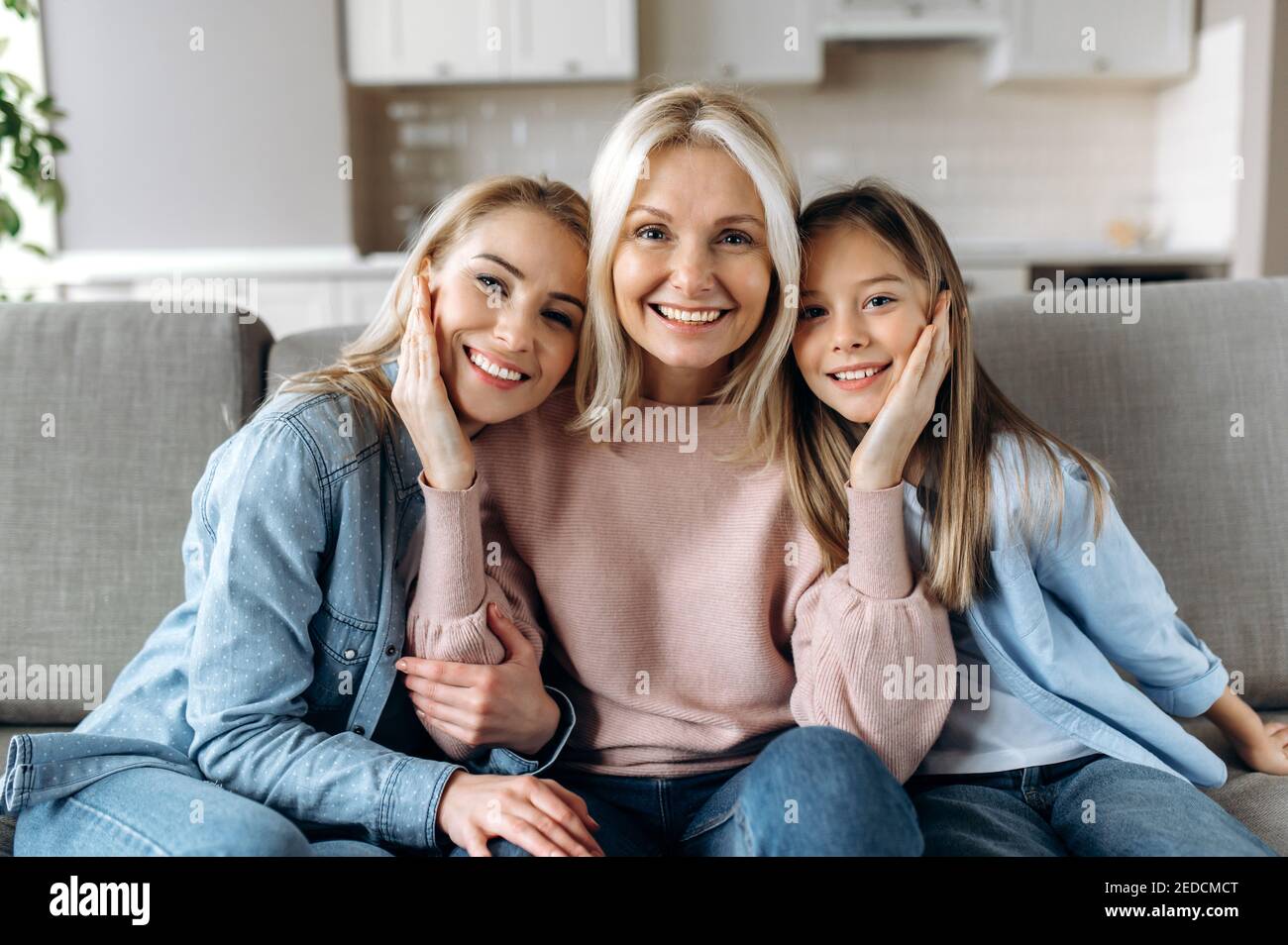 Ritratto di una felice nonna caucasica, figlia e nipote, sono seduti a casa sul divano, guardando la macchina fotografica e sorridendo. Rapporti e valori familiari Foto Stock