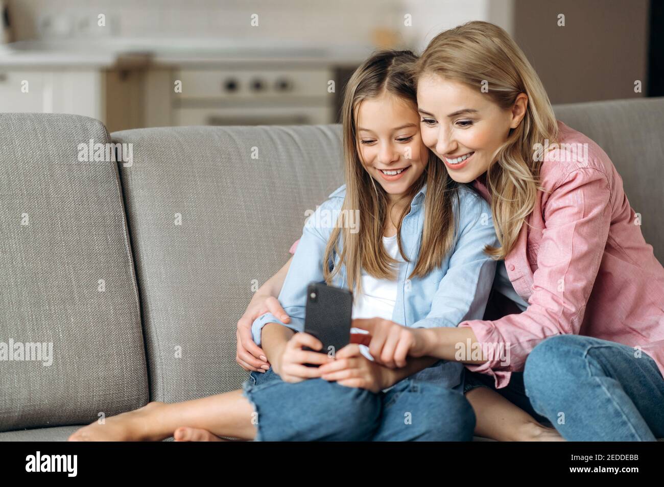 Sorridente mamma e figlia bionda felici vestite di abiti casual eleganti, rilassarsi a casa, utilizzare il telefono, navigare in Internet, social network, prendere un selfie Foto Stock