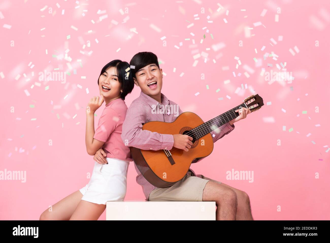 Felice giovane asiatico che suona la chitarra e canta con lui studio di fidanzata girato su sfondo rosa Foto Stock