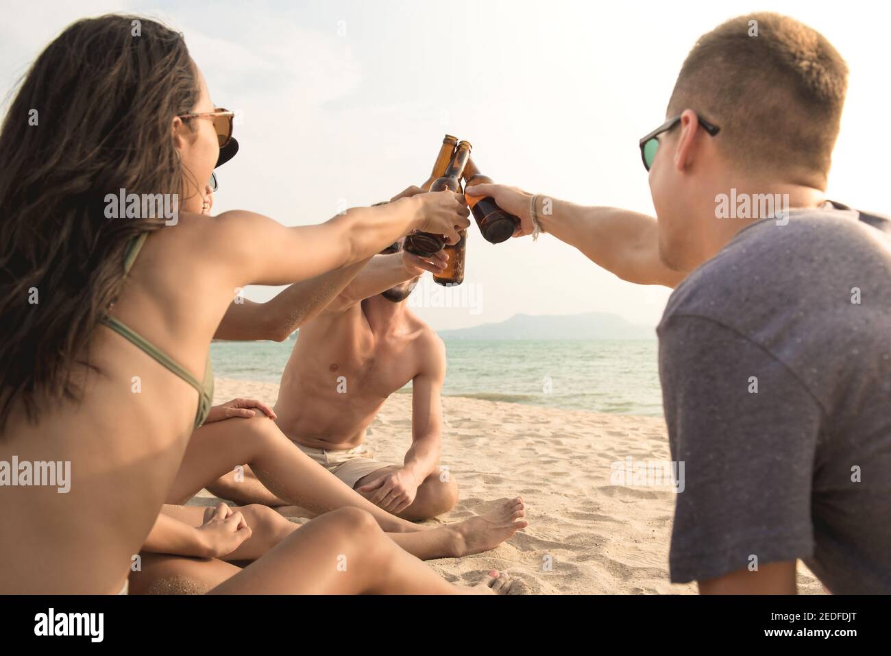 Gruppo di amici che hanno partito che claging bottiglie di birra che fanno un brindisi prima di bere in spiaggia in estate Foto Stock