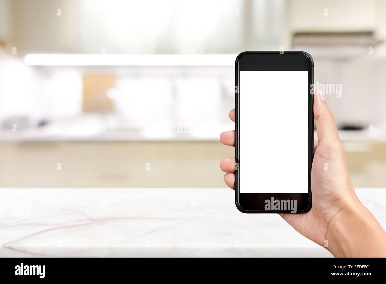 Mano che tiene lo smartphone con schermo vuoto su sfondo di cucina sfocato Foto Stock
