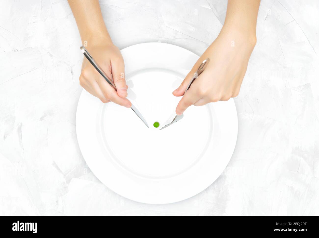 Le mani femminili cercano di catturare un piccolo pisello verde su un grande piatto bianco con coltello e forchetta. Concetto di disturbo alimentare. Foto Stock