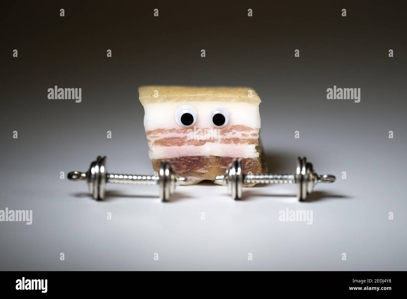 Personaggio divertente fatto di grasso di maiale salato guardando pesanti manubri su uno sfondo neutro. Concetto creativo di workout di perdita di peso e bruciatura del grasso. Foto Stock