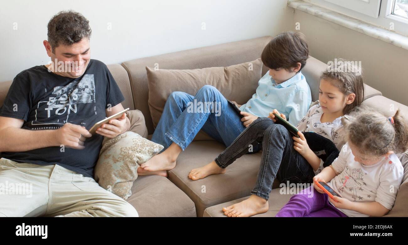 padre e i suoi figli sono seduti sul divano. Giocare a giochi su internet, hanno tutti telefoni cellulari e tablet digitali nelle loro mani Foto Stock