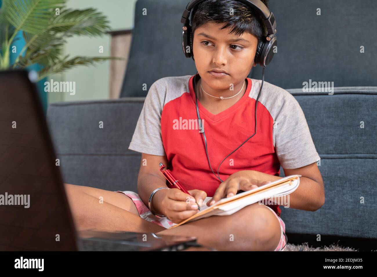 Un bambino serio con le cuffie che annotano fino a prenotare vedendo il laptop durante la classe in linea a casa - concetto di classe in linea, formazione in linea Foto Stock