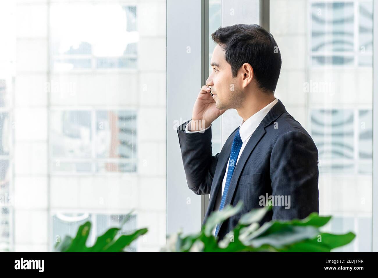 Uomo asiatico in tuta da lavoro formale che chiama sul telefono cellulare e guardando verso l'esterno la finestra di vetro dell'ufficio Foto Stock