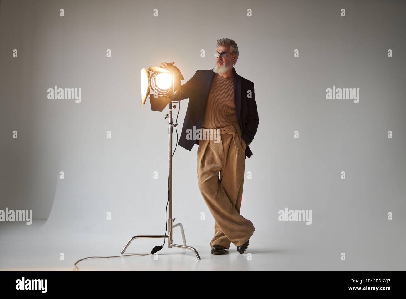 Foto a tutta lunghezza di un elegante uomo di mezza età in occhiali da vista, in piedi accanto al riflettore dello studio mentre si posa per la fotocamera su sfondo bianco. Foto di moda, concetto di stile Foto Stock