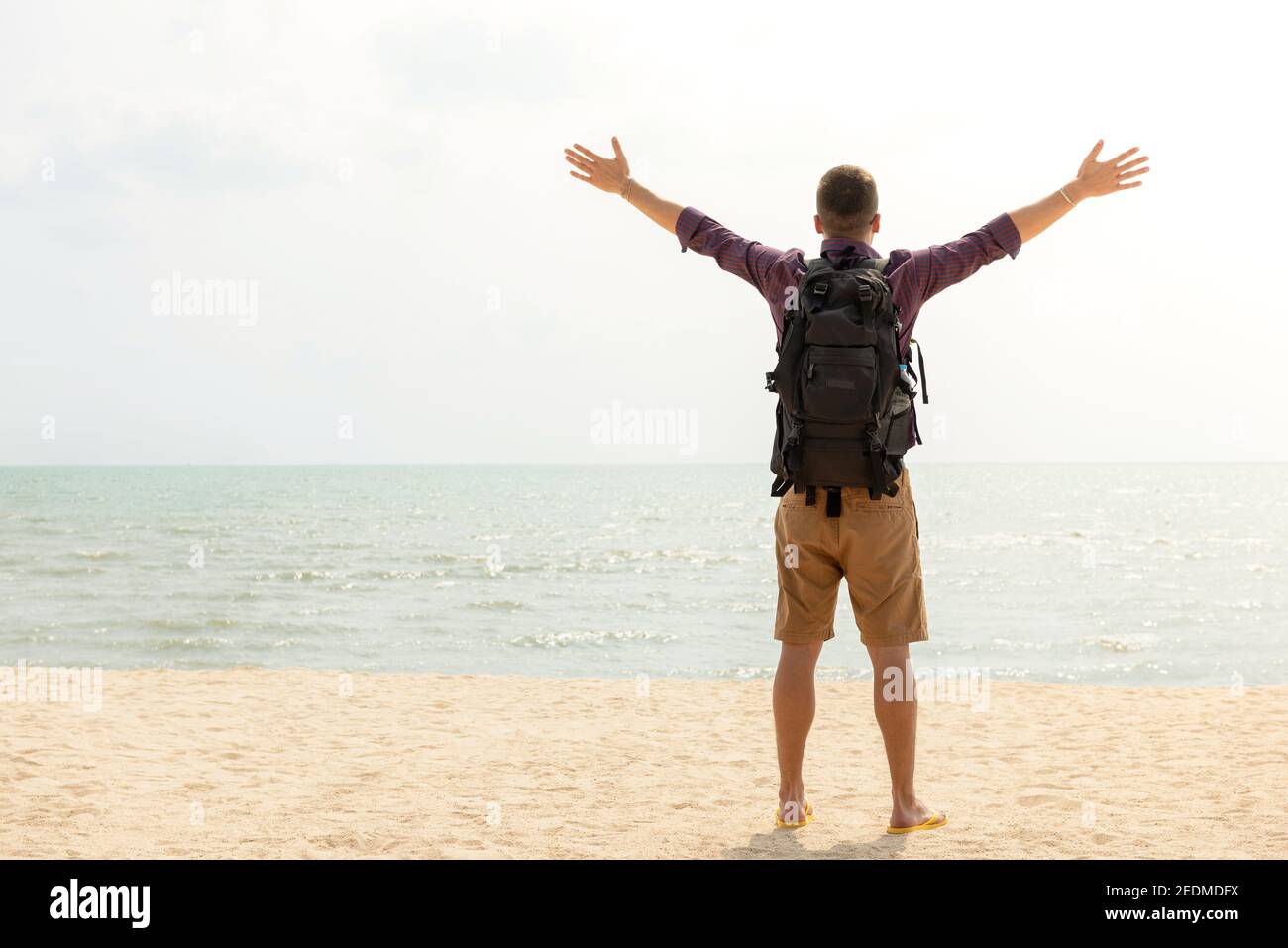 Felice turista zaino in spalla con braccia aperte in spiaggia nelle vacanze estive, vista sul retro con spazio copia Foto Stock