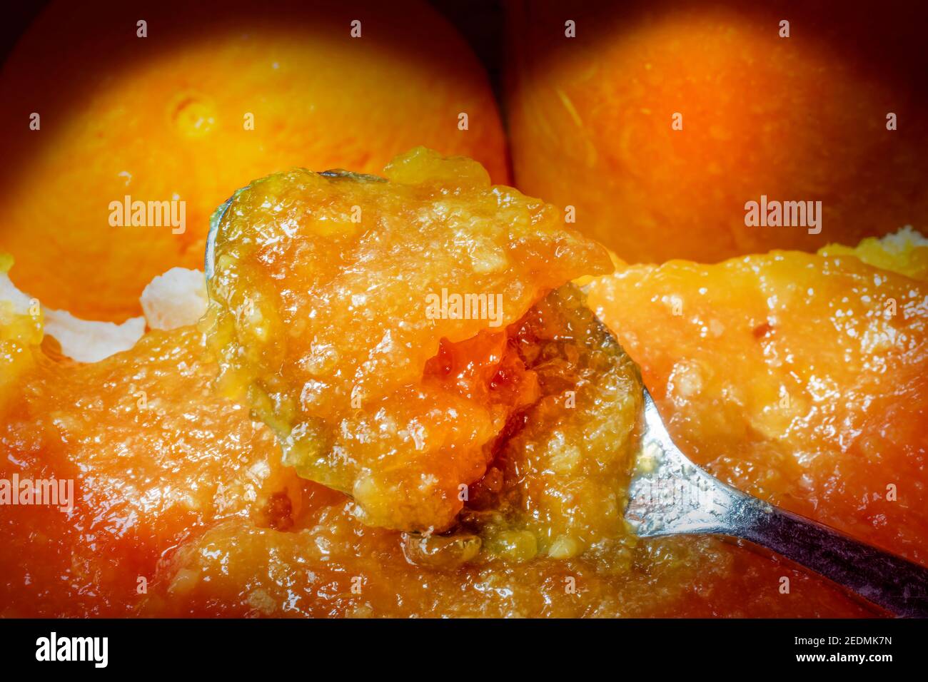 Cucchiaio annegato in marmellata d'arancia fresca. Spalmare su un pezzo di pane. Parte di arancione e vaso come sfondo Foto Stock
