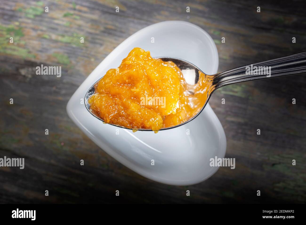 Cucchiaio di marmellata d'arancia fresca . Sotto c'è una ciotola a forma di cuore bianco su sfondo di legno. Foto Stock