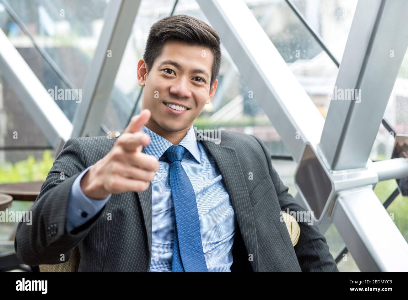 Cordiale e simpatico giovane uomo d'affari asiatico seduto nella sala d'ufficio, sorridente e rivolto verso la macchina fotografica Foto Stock