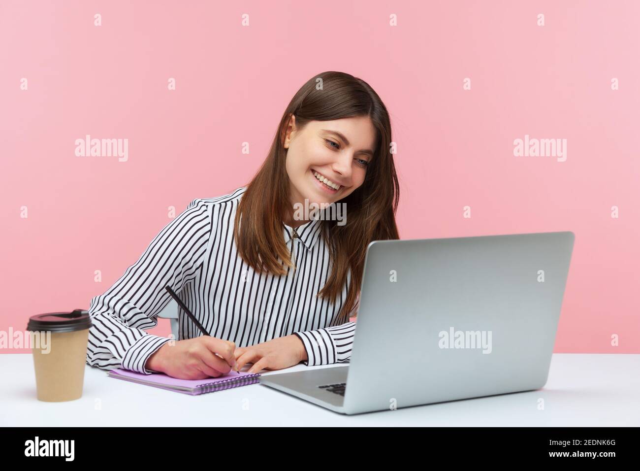 Sorridente lavoratore positivo in ufficio donna con camicia a righe che prende appunti con la matita al blocco note guardando il display del computer portatile, ha videochiamata conferenza. Interno Foto Stock