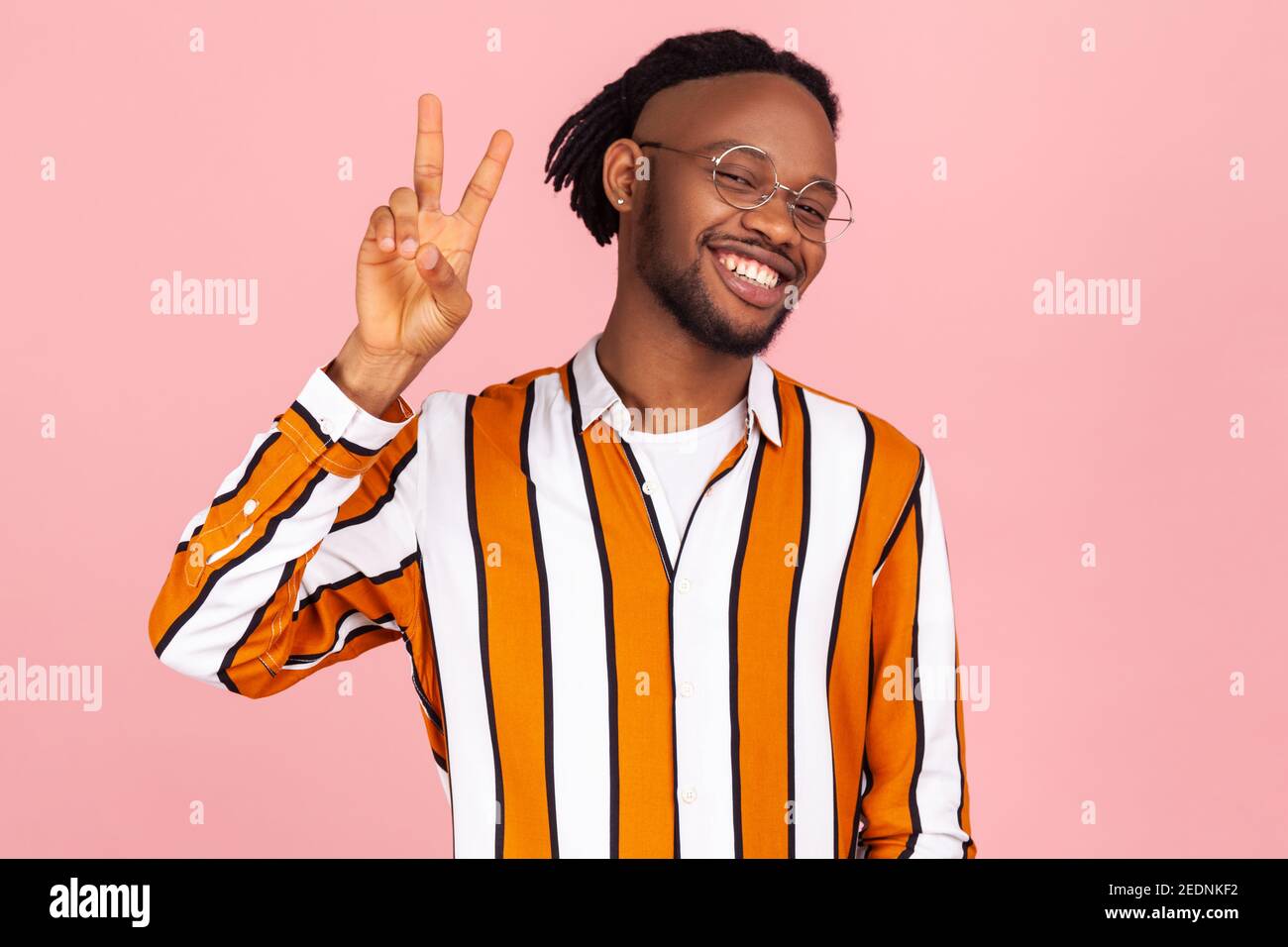 Eccitato allegro afro-americano uomo con barba e dreadlock in occhiali e elegante camicia a righe che mostra segno di pace, v gesto e sorridente. Interno Foto Stock