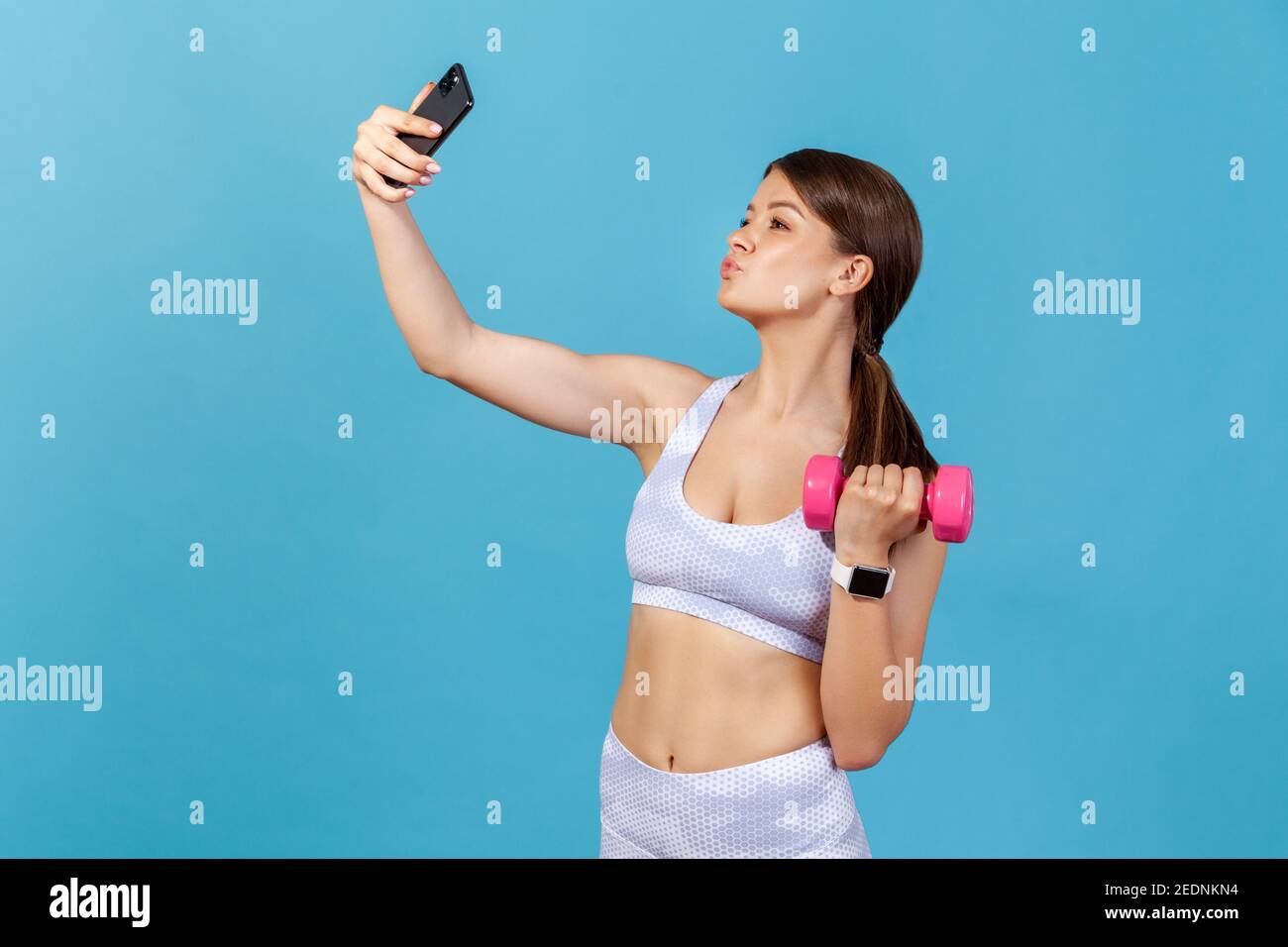 Profilo ritratto sottile blogger donna in bianco sportswear in posa sulla fotocamera del suo smartphone, la registrazione di allenamento con dumbbell tutorial. Studio interno sh Foto Stock