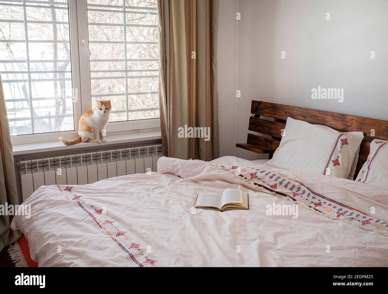 Interno della camera da letto. Biancheria da letto leggera, libro sul letto e gatto sulla finestra. Foto Stock