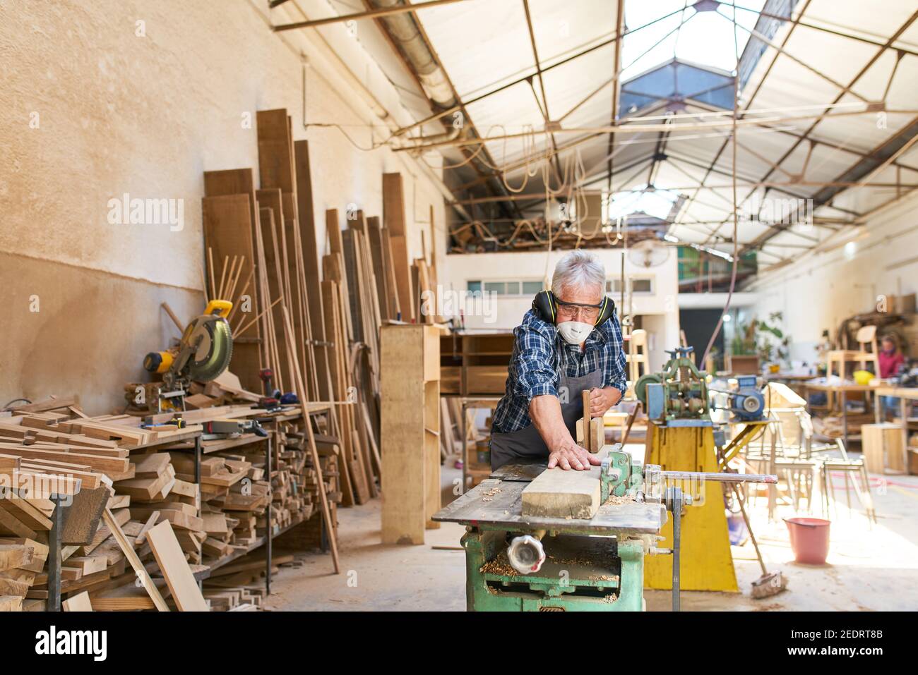 Anziano artigiano nel laboratorio di Falegnameria mentre piallando legno con una maschera e una protezione per l'udito Foto Stock