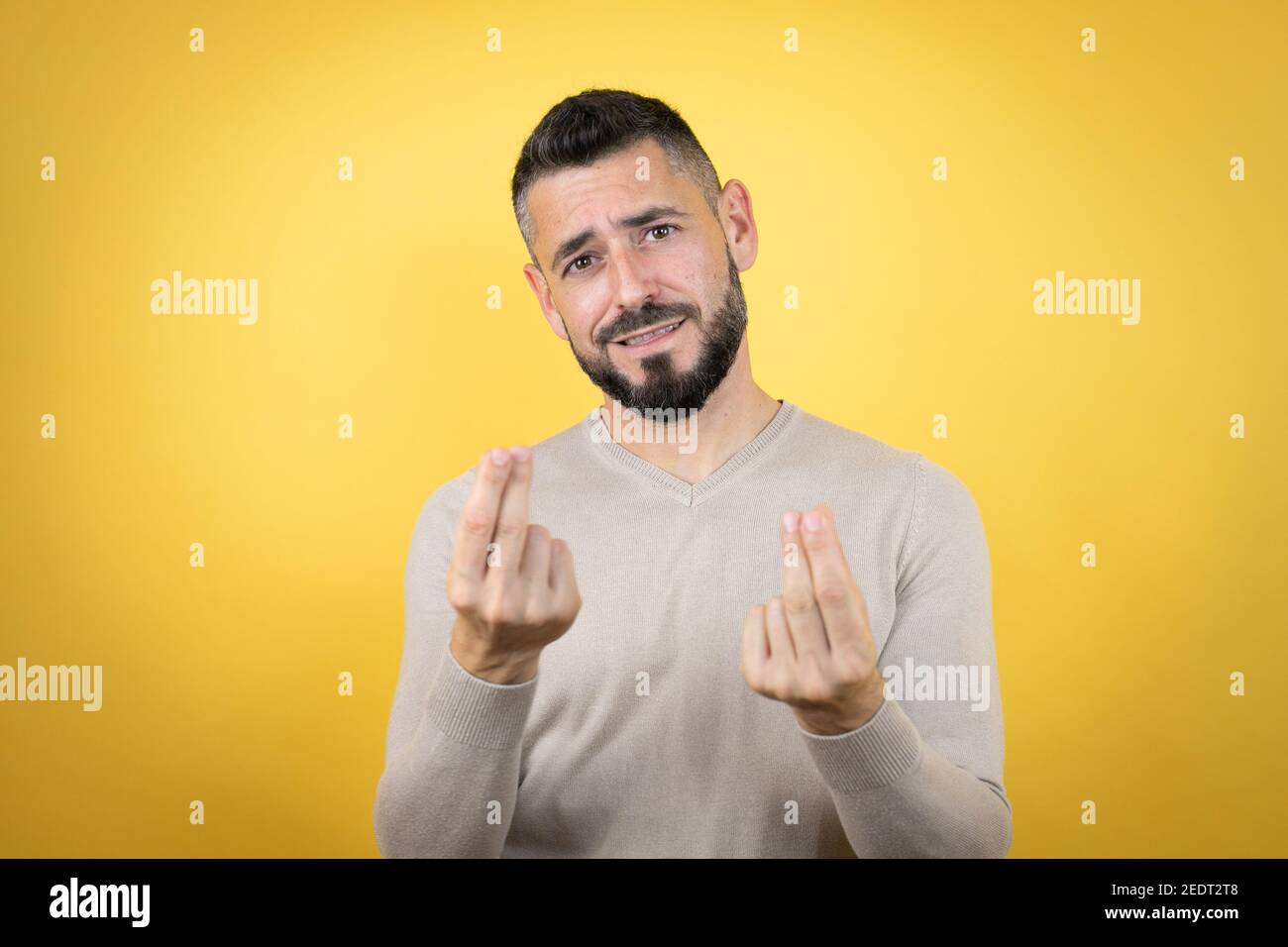Bell'uomo con barba che indossa maglione su sfondo giallo facendo gesti di denaro con le mani, chiedendo il pagamento dello stipendio, milionario business Foto Stock