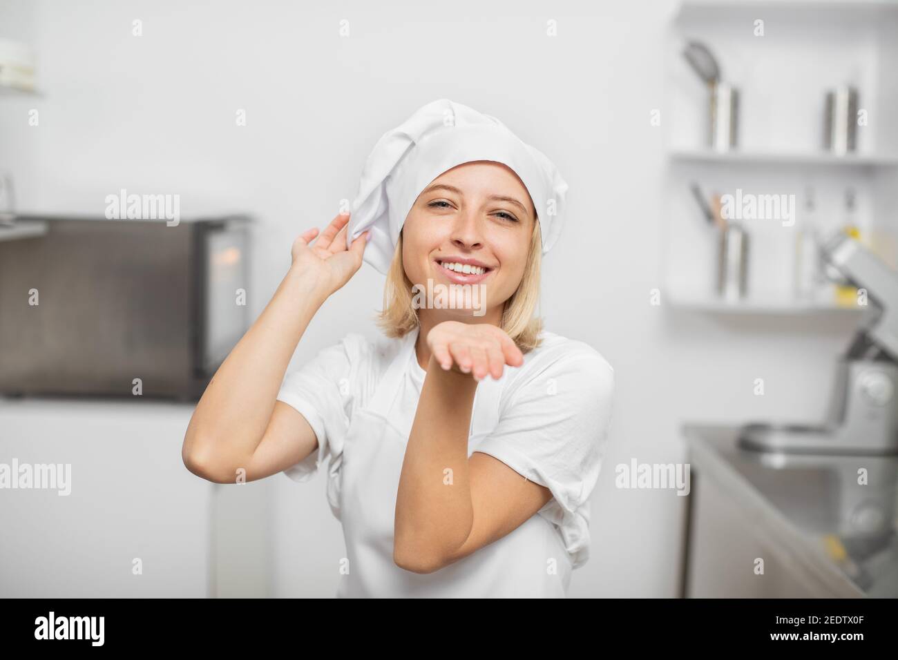 Persone, occupazioni e concetto di lavoro. Giovane bella donna confettatrice, indossando grembiule e cappello bianco, inviando bacio aria alla macchina fotografica con sorriso Foto Stock