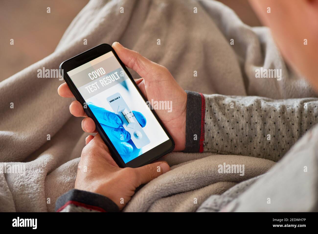 primo piano di un giovane caucasico in pigiama, seduto comodamente su un divano avvolto in una coperta grigia, controllando il risultato del suo covid test in un simul Foto Stock