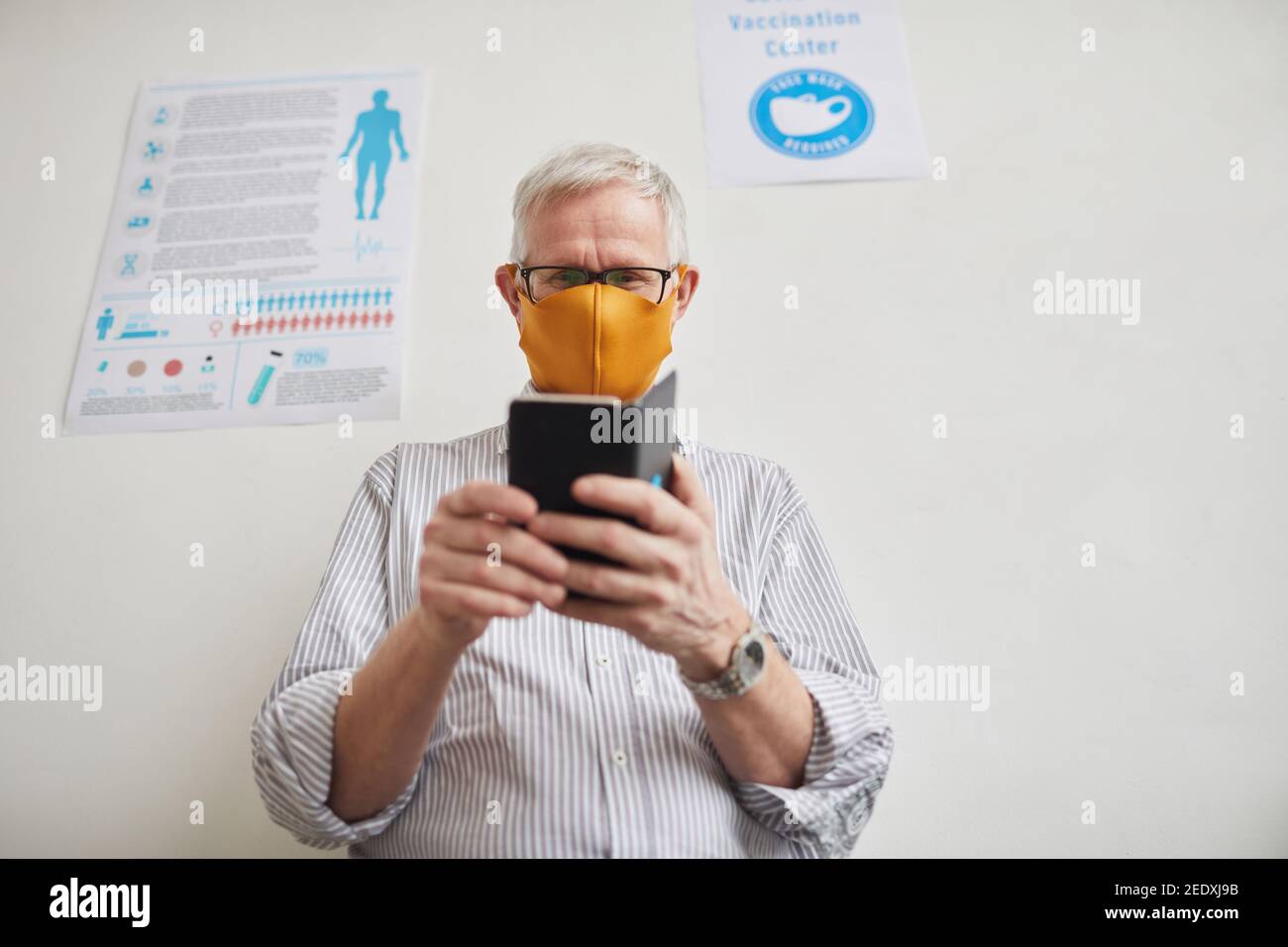 Ritratto ad angolo basso di un uomo anziano con capelli bianchi che usa uno smartphone mentre si attende in fila presso la clinica medica, spazio copia Foto Stock