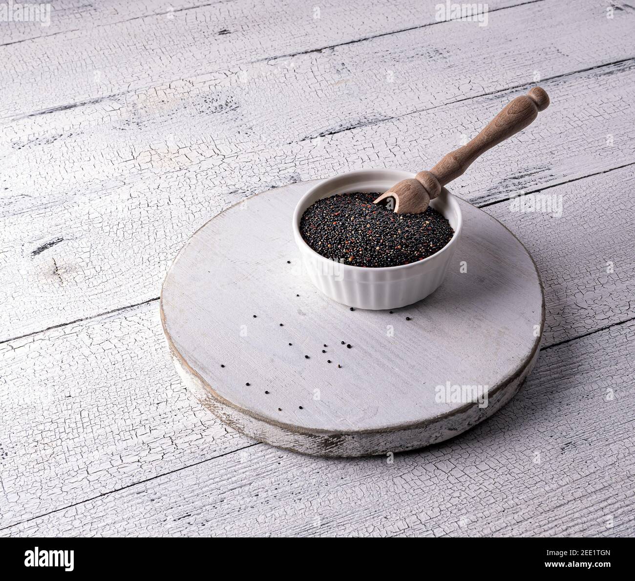 Semi di quinoa crudo nero non cotti e scoop in ciotola bianca a bordo e su tavolo di legno bianco. Pochi semi sono sparsi intorno. Cibo eccellente con copia grande Foto Stock